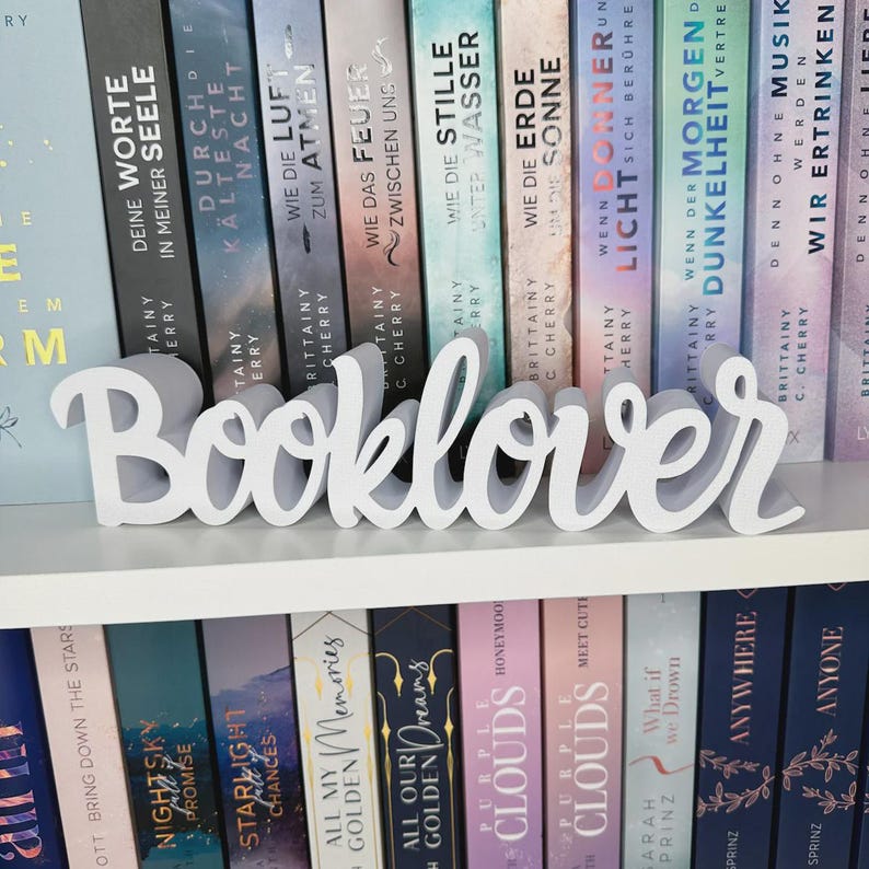 Op de afbeelding: Een wit "Booklover" bord in cursiefschrift staat op een witte plank voor een rij kleurrijke boeken. De boeken hebben Duitse titels. Het bord is een decoratief item voor boekenliefhebbers.