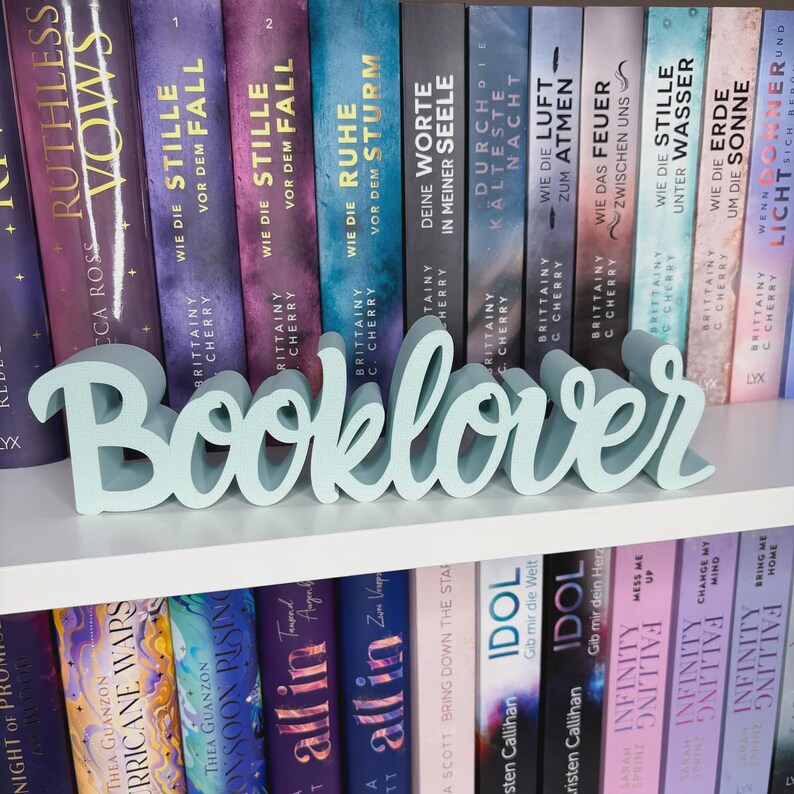 Op de afbeelding: Een lichtblauw houten "Booklover" bord staat op een witte plank voor een rij boeken. De boeken hebben kleurrijke covers met titels in het Engels en Duits. Het bord is in een cursief lettertype.