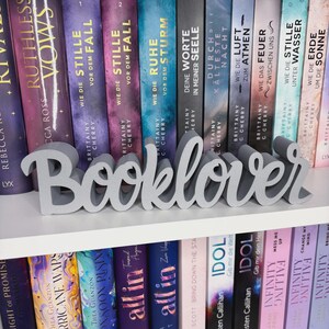Op de afbeelding: Een grijze "Booklover" woordkunst decoratie op een witte plank voor een rij boeken. De boeken hebben kleurrijke covers met titels in het Engels en Duits. Een decoratief item voor boekenliefhebbers.