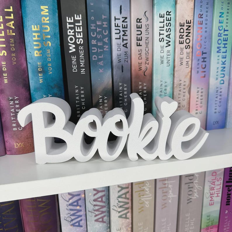 Op de afbeelding: Een witte 'Bookie' woordkunst siert een witte plank voor een rij boeken met Duitse titels. Het woord is in een cursief lettertype. De boeken hebben kleurrijke ruggen. Een decoratief item voor boekenliefhebbers.