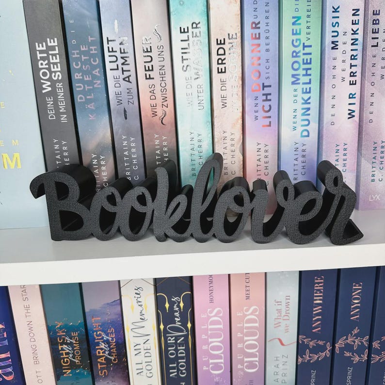 Op de afbeelding: Een donkergrijs "Booklover" woordkunst decorstuk, weergegeven op een witte plank voor een rij kleurrijke boeken. De boeken hebben Duitse titels. De woordkunst is in een cursief lettertype.