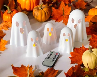 Pode incluir: Um conjunto de seis luzes LED brancas em forma de fantasma com um controle remoto. As luzes estão dispostas em uma superfície branca com folhas de outono e abóboras.