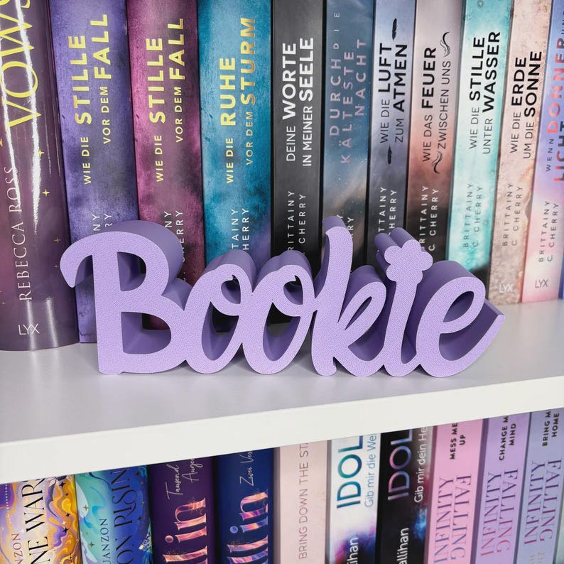 Op de afbeelding: Een lavendelkleurige "Bookie" woordkunst siert een witte plank, met een verzameling boeken op de achtergrond. De boeken hebben kleurrijke ruggen met Duitse titels. Het woord "Bookie" is in cursief.