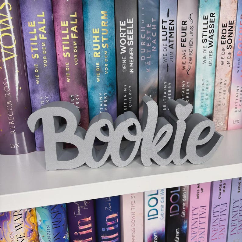 Op de afbeelding: Een grijs 'Bookie' woordkunst decorstuk staat op een witte plank voor een rij kleurrijke boeken. De boeken hebben Duitse titels. Het woord 'Bookie' is in een cursief lettertype.