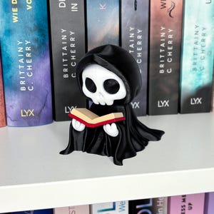 Könnte beinhalten: Eine schwarz-weiße Figur des Sensenmanns, der ein Buch mit rotem Einband liest. Der Sensenmann trägt eine schwarze Kapuzenrobe. Die Figur steht auf einem weißen Regal vor einer Reihe von Büchern.