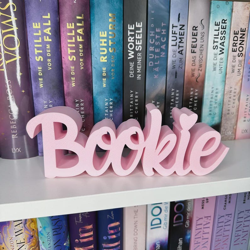 Op de afbeelding: Een roze "Bookie" woordteken, weergegeven op een witte plank voor een verzameling boeken. Het teken is in een cursief lettertype en is een decoratief item voor boekenliefhebbers. De boeken hebben kleurrijke ruggen.