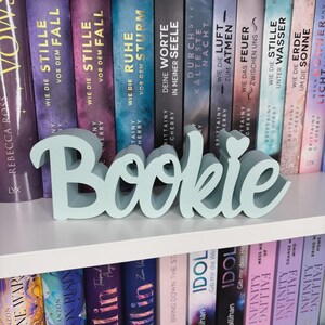 Op de afbeelding: Een lichtblauwe "Bookie" woordkunst decoratie, tentoongesteld op een witte plank voor een rij boeken. Het woord is in een cursief lettertype met een hartvormige stip boven de "i". De boeken hebben kleurrijke ruggen.