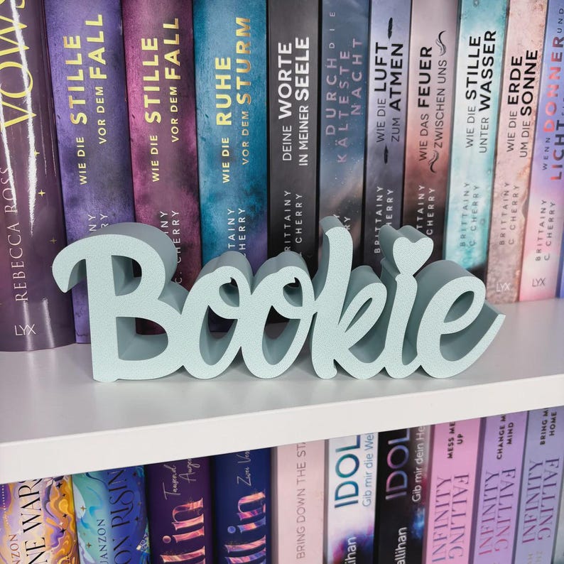 Op de afbeelding: Een lichtblauwe "Bookie" woordkunst decoratie, tentoongesteld op een witte plank voor een rij boeken. Het woord is in een cursief lettertype met een hartvormige stip boven de "i". De boeken hebben kleurrijke ruggen.