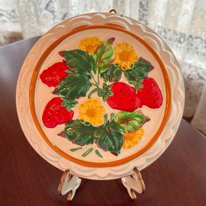 May include: A white ceramic plate with a raised design of red strawberries and yellow flowers. The plate has a scalloped edge and is on a stand.