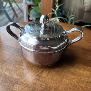 以下が含まれることがあります： 蓋と2つのハンドルが付いた銀色の砂糖入れ。ボウルは丸く、木製の表面に置かれています。蓋の上部には小さなノブが付いています。