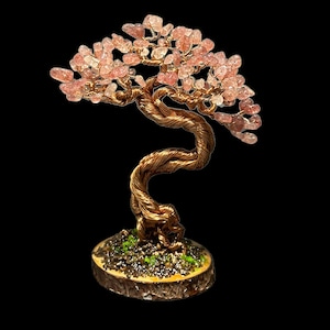 May include: A pink quartz wire wrapped tree with a twisted copper trunk and a wooden base. The tree is decorated with small pink quartz crystals.