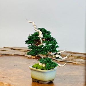 May include: A miniature wire bonsai tree with green leaves and silver branches in a gray ceramic pot. The bonsai is sitting on a wooden surface.