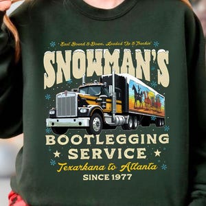 May include: Dark green sweatshirt with a graphic of a semi-truck. The text on the sweatshirt reads "SNOWMAN'S BOOTLEGGING SERVICE Texarkana to Atlanta SINCE 1977" with snowflakes and stars. The truck has a yellow and black cab and a trailer with a horse painting.