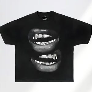 May include: Black t-shirt featuring a grayscale image of two sets of lips with silver grillz. The design is a close-up, highlighting the teeth and the glossy texture of the lips. The shirt is a classic crew neck style.