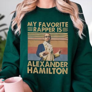 A person wearing a green sweatshirt with a graphic design featuring a man in a suit and the text "My favorite rapper is Alexander Hamilton" printed on it.