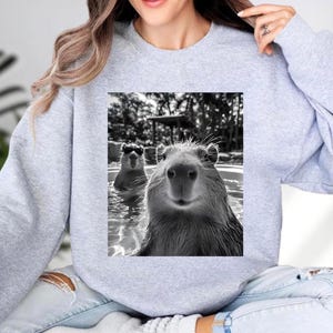 A woman wearing a gray sweatshirt with a black and white photograph of two beavers swimming in a pool.