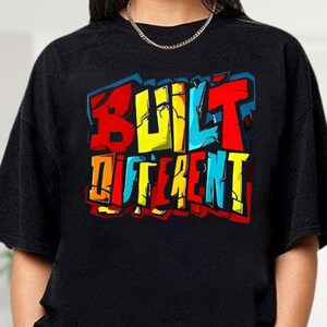 May include: Black sweatshirt with the words "BUILT DIFFERENT" in a colorful graffiti-style design. The text features red, yellow, blue, and orange colors with a white outline. The sweatshirt has a crew neck and long sleeves.