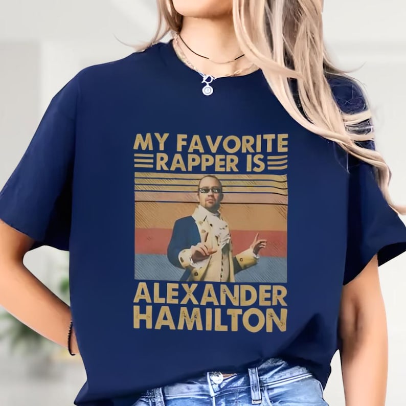 a blue t-shirt with a graphic design of a person wearing a suit and tie, along with text that says "My favorite rapper is Alexander Hamilton".