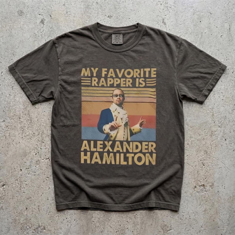 A t-shirt with a graphic design featuring a man in a suit and tie, along with text that says "My favorite rapper is Alexander Hamilton".