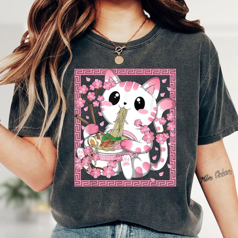 May include: Dark gray t-shirt featuring a cute cartoon cat with pink accents eating ramen from a bowl. The cat is surrounded by pink cherry blossoms and a pink decorative border. The design is centered on the shirt.