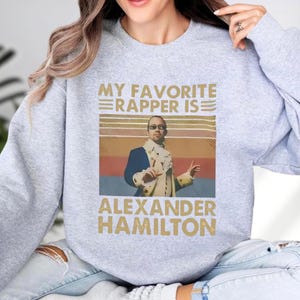 A gray sweatshirt with a graphic design featuring a man in a historical military uniform and the text "My favorite rap artist is Alexander Hamilton" printed on it.