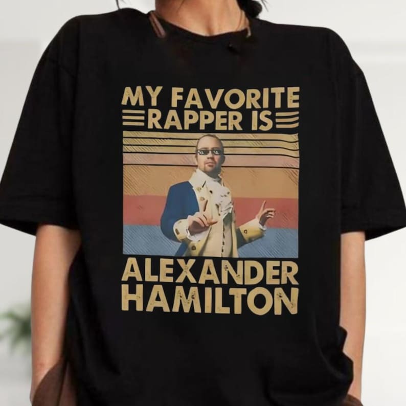 a person wearing a black t-shirt with a graphic design that says "My favorite rapper is Alexander Hamilton" and includes a portrait of Alexander Hamilton.