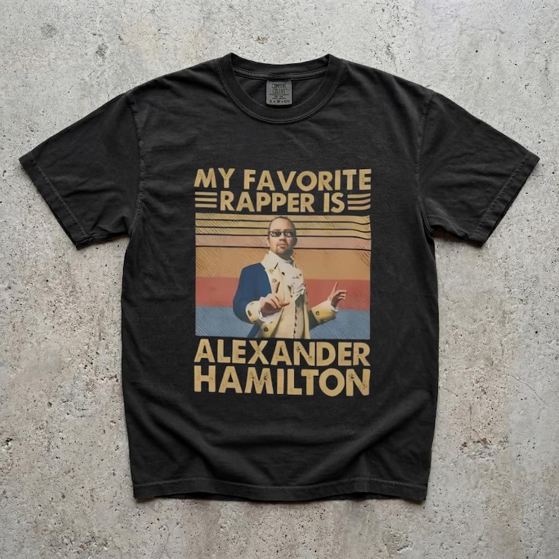 A black t-shirt with a graphic design featuring a person wearing a suit and tie, along with text that says "My favorite rapper is Alexander Hamilton".
