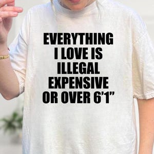 May include: Cream-colored t-shirt with the text "EVERYTHING I LOVE IS ILLEGAL EXPENSIVE OR OVER 6'1"" in bold black letters. The shirt has short sleeves and a crew neck.