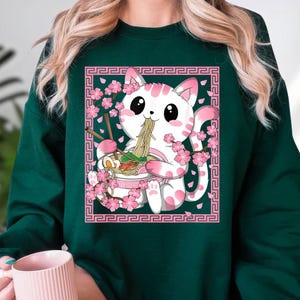 May include: Dark green sweatshirt featuring a white cat with pink spots, eating ramen from a bowl. The cat is surrounded by pink cherry blossoms and a pink decorative border. The design is a cute, cartoon style.