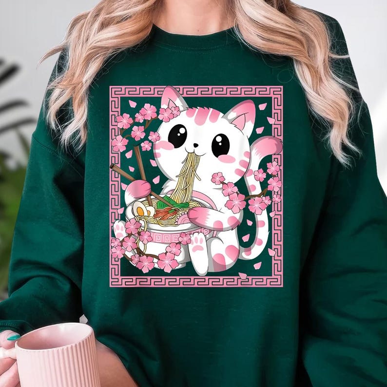 May include: Dark green sweatshirt featuring a white cat with pink spots, eating ramen from a bowl. The cat is surrounded by pink cherry blossoms and a pink decorative border. The design is a cute, cartoon style.