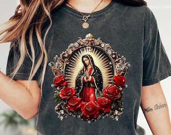May include: A charcoal gray short-sleeve t-shirt featuring a central graphic of the Virgin of Guadalupe. The figure is framed by an ornate gold oval border with sparkling accents, surrounded by vibrant red roses and golden rays.