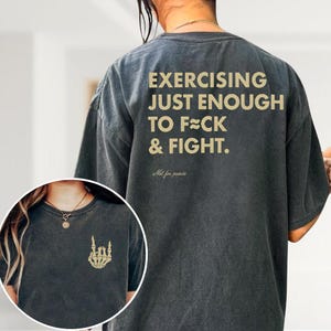 May include: Dark gray t-shirt with the text "EXERCISING JUST ENOUGH TO F**K & FIGHT" in gold lettering on the back. A smaller image on the front shows a gold skeleton hand. The shirt is a casual style.