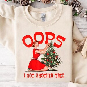 May include: Cream-colored sweatshirt with a retro graphic. The graphic features the word "OOPS" in red above a woman in a red dress decorating a Christmas tree. The text "I GOT ANOTHER TREE" is below the image in red.