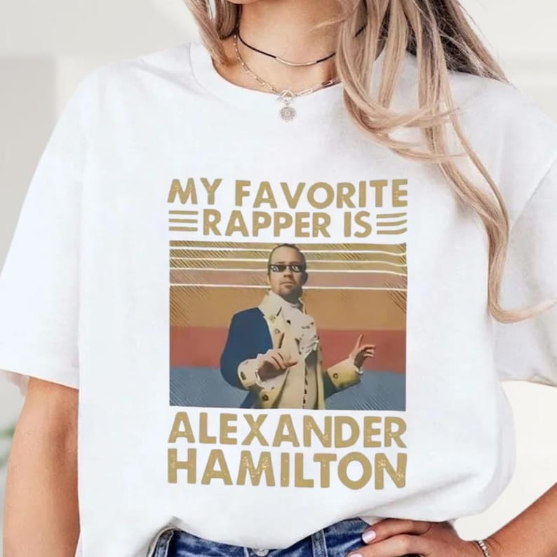 A white t-shirt with a graphic design featuring a man wearing a suit and tie, along with text that says "My favorite rapper is Alexander Hamilton".