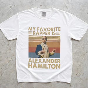 A white t-shirt with a graphic design featuring a man in a suit and sunglasses, along with text that says "My favorite rapper is Alexander Hamilton".