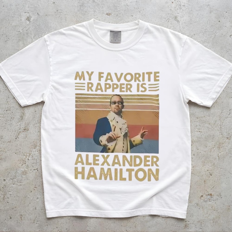 A white t-shirt with a graphic design featuring a man in a suit and sunglasses, along with text that says "My favorite rapper is Alexander Hamilton".