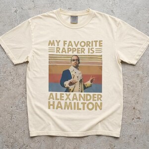 A white t-shirt with a graphic design featuring a man in a suit and tie, along with text that says "My favorite rapper is Alexander Hamilton".