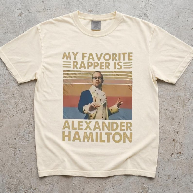 A white t-shirt with a graphic design featuring a man in a suit and tie, along with text that says "My favorite rapper is Alexander Hamilton".