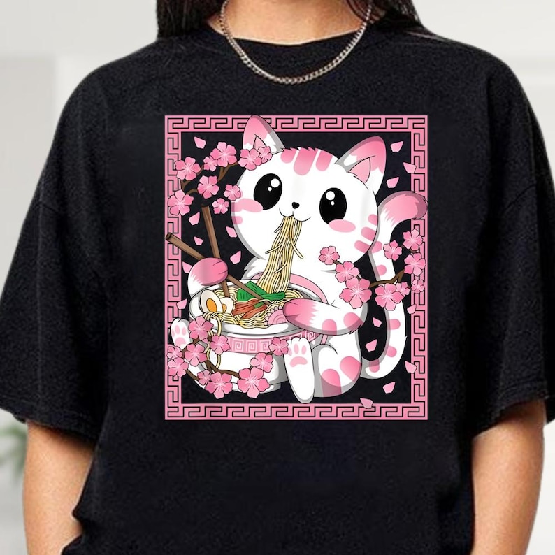 May include: Black sweatshirt featuring a white and pink cat enjoying ramen. The cat is surrounded by pink cherry blossoms and holds chopsticks. The design is framed by a pink border with a repeating pattern.