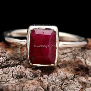 May include: A silver ring featuring a rectangular, deep red gemstone. The gemstone is set in a bezel setting, and the band is thin and polished. The ring is displayed on a textured, brown surface against a black background.
