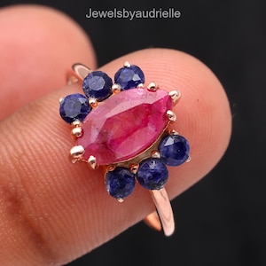 May include: A rose gold ring with a central marquise-cut ruby, encircled by dark blue sapphires. The ring has a floral design, with the gemstones set in a delicate pattern. The ring is shown against a dark backdrop. The ring is a piece of jewellery.