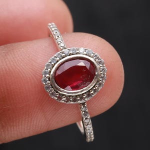 May include: A silver ring featuring a large, oval-cut, deep red gemstone, surrounded by smaller, clear stones. The ring's band is also adorned with small, clear stones, creating a sparkling effect. The ring is held against a dark background.