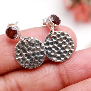 May include: Silver stud earrings with a hammered disc design and a small, round, red gemstone. The earrings are shown against a light background. The earrings are a jewelry item.
