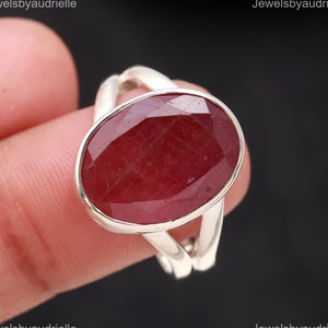 May include: A silver ring featuring a large, oval-cut ruby gemstone. The ruby is a deep red color and is set in a simple, elegant silver band. The ring is shown on a finger against a dark background.