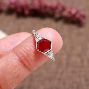 May include: A silver ring featuring a hexagonal, deep red gemstone. The ring is accented with small, clear gemstones on either side of the central stone. The ring is shown on a finger against a blurred background.
