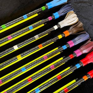 May include: A collection of eight long, slender rods with black wrapped handles and bright yellow shafts. Each rod features a colourful leather-like fringe at the top, secured by a matching coloured band, with additional accent tape along the handle.