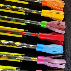 May include: A collection of eight riding crops featuring bright yellow shafts wrapped in black leather strips with metallic underlays. Each crop ends in a colourful fringed leather handle in blue, orange, yellow, maroon, red, light blue, purple, and brown.