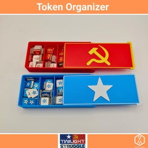 May include: Two token organizers, one red and one blue, with lids featuring a hammer and sickle and a white star. The organizers are filled with game tokens. The text "Token Organizer" is at the top.