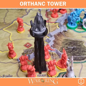 May include: A black plastic Orthanc tower miniature featuring a grey figure of Saruman on top, positioned on a board game map. The scene includes several red and blue plastic soldier miniatures placed around the tower on the printed game board.