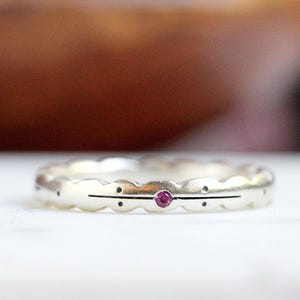 May include: A silver ring with a scalloped edge design. The ring features a small, round, red gemstone set in the center, with a horizontal line and dots on either side. The ring is displayed on a white surface.